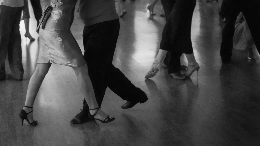 Black and white close-up of several dancers' legs and feet moving across a wooden social dance floor